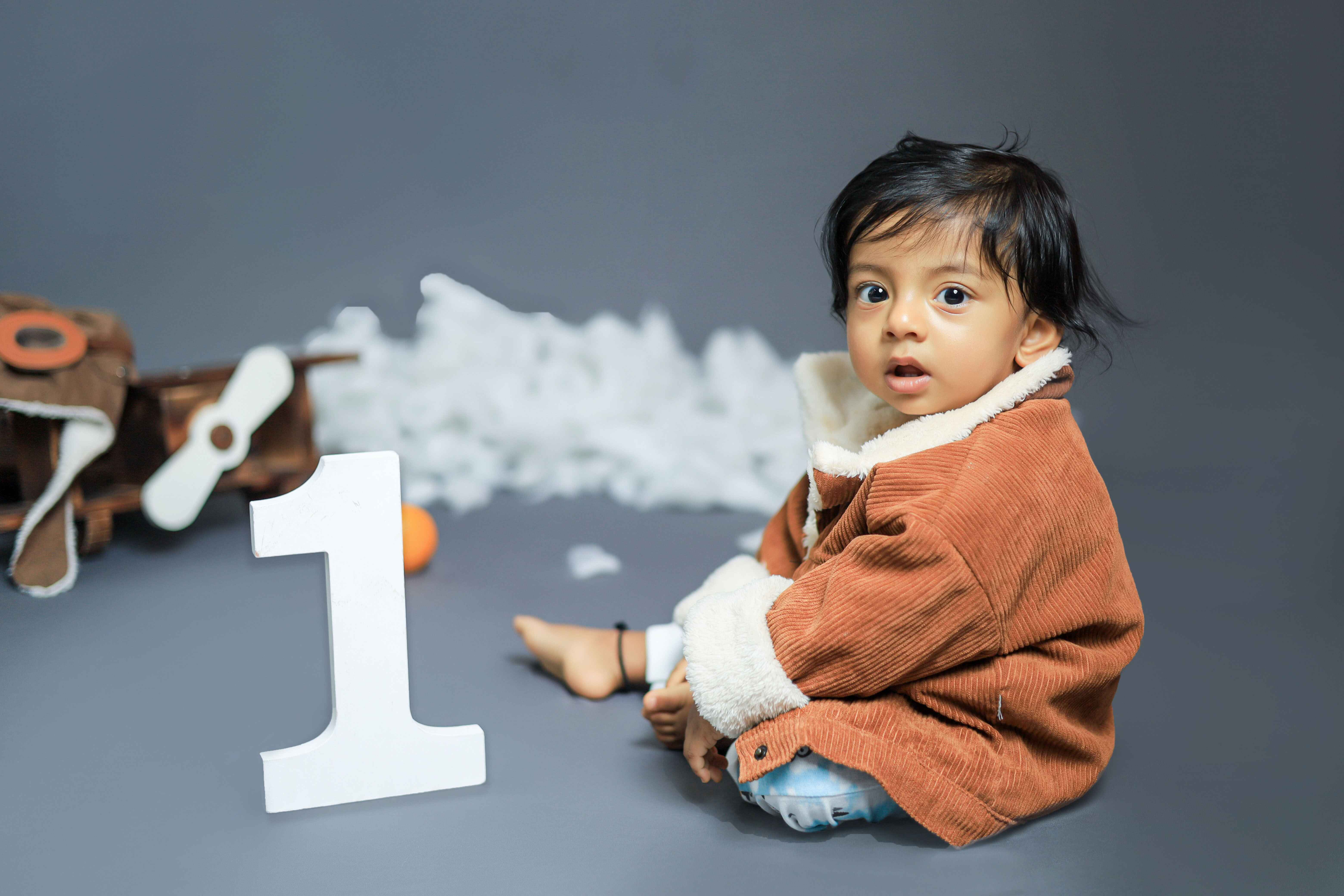 A baby in a brown jacket with a one-year prop, captured in a cheerful and adorable baby photoshoot.